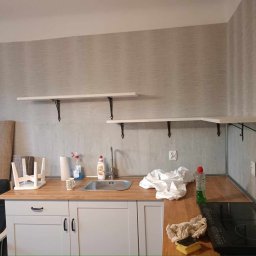 USŁUGI BUDOWLANE PAWEŁ LAUTER - Kuchnia w trakcie sprzątania po remoncie: blat z zlewozmywakiem, płyta indukcyjna, wiszące półki, środki czystości i ściereczki. Widoczne ślady prac wykończeniowych.