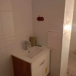 Umywalka z szafką w trakcie montażu w łazience wykończonej białymi, prostokątnymi płytkami; widoczne otwarte gniazdko elektryczne, wałek do malowania i ołówek.