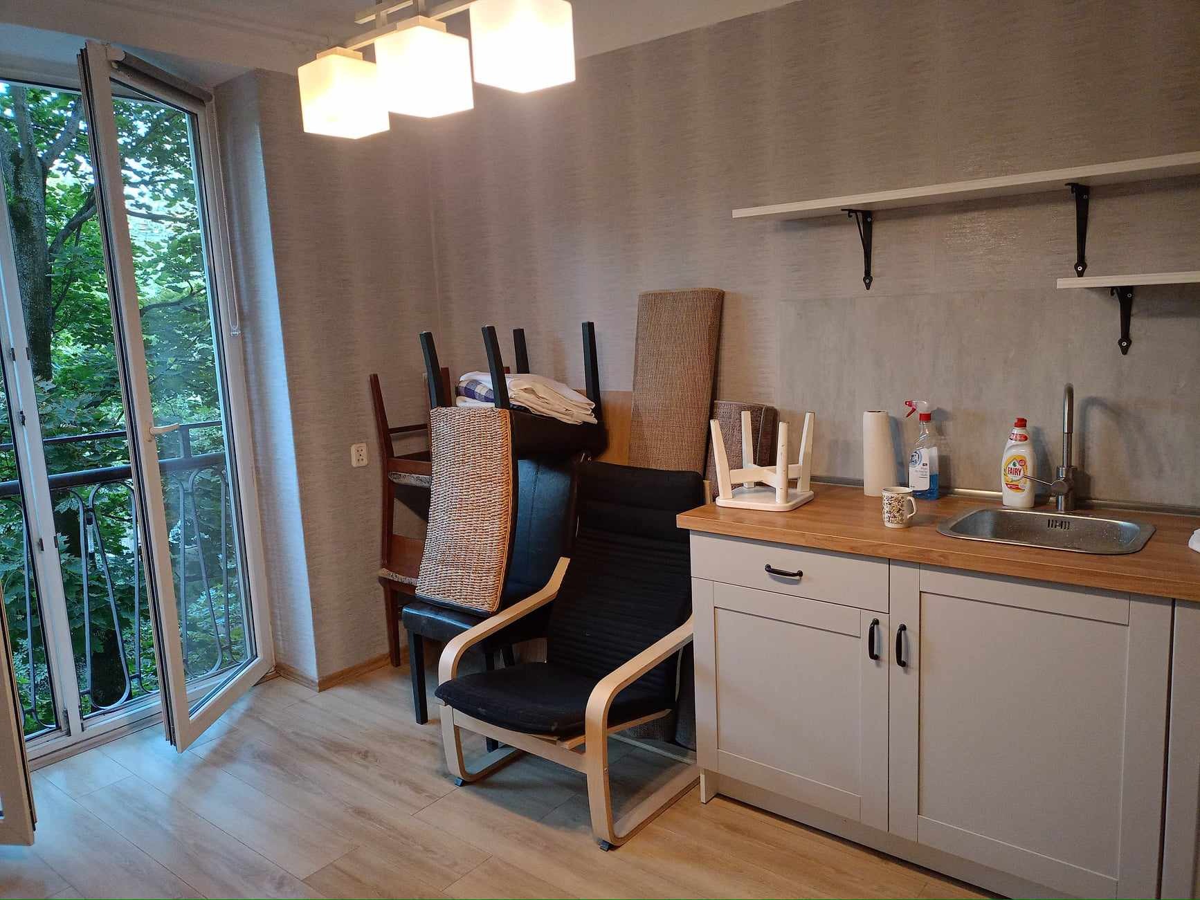 Wnętrze pokoju z otwartym balkonem, widoczna zabudowa kuchenna z drewnianym blatem, krzesła ustawione obok, na podłodze panele imitujące drewno.