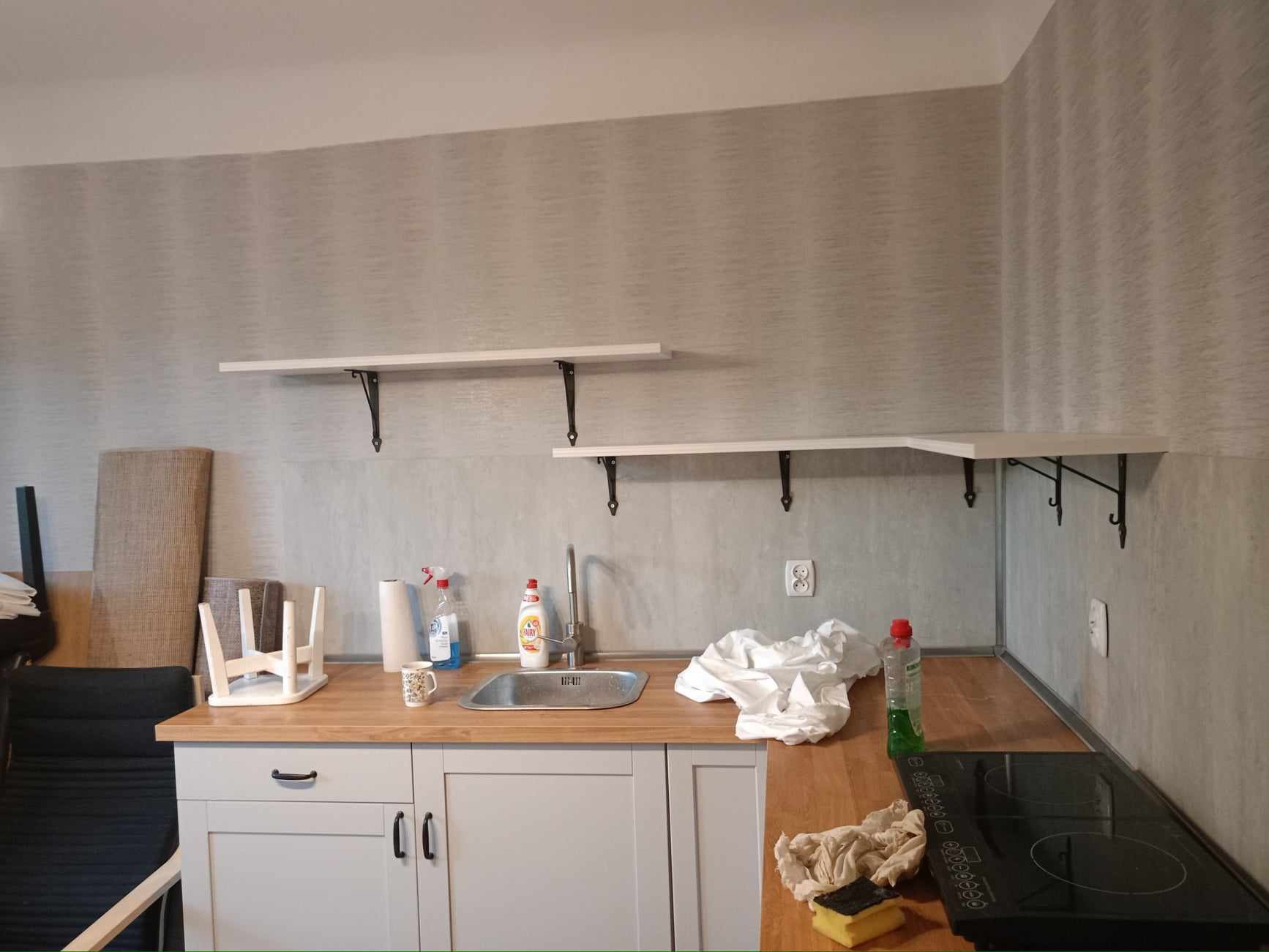 Kuchnia w trakcie sprzątania po remoncie: blat z zlewozmywakiem, płyta indukcyjna, wiszące półki, środki czystości i ściereczki. Widoczne ślady prac wykończeniowych.