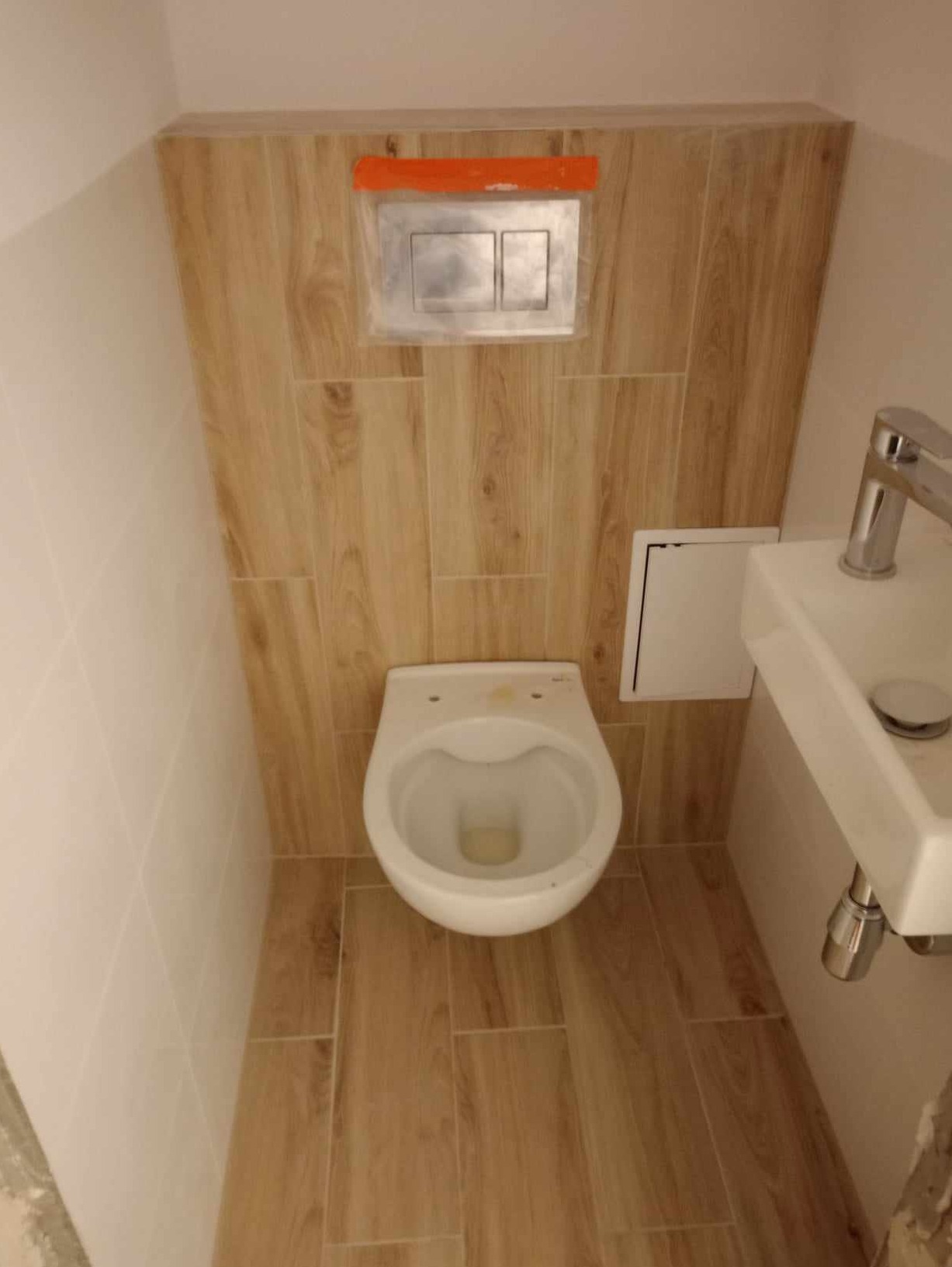Zawieszona miska WC i umywalka w małej łazience wykończonej płytkami imitującymi drewno, z widocznym przyciskiem spłukującym zabezpieczonym folią.