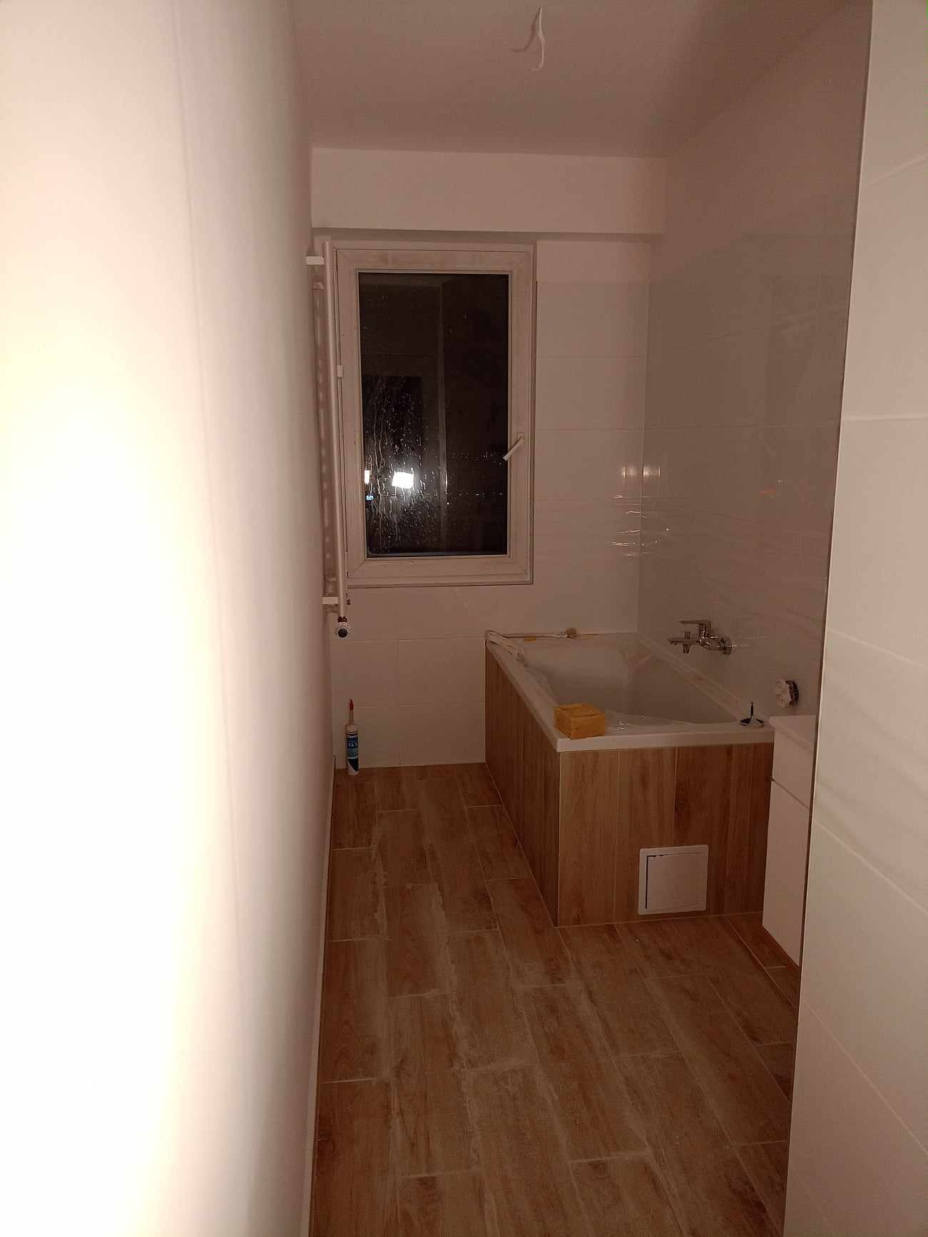 Nowoczesna łazienka po remoncie: białe płytki ścienne, drewnopodobna obudowa wanny, podłoga z płytek imitujących drewno, okno z widokiem na nocne miasto, tuba silikonu przy ścianie.