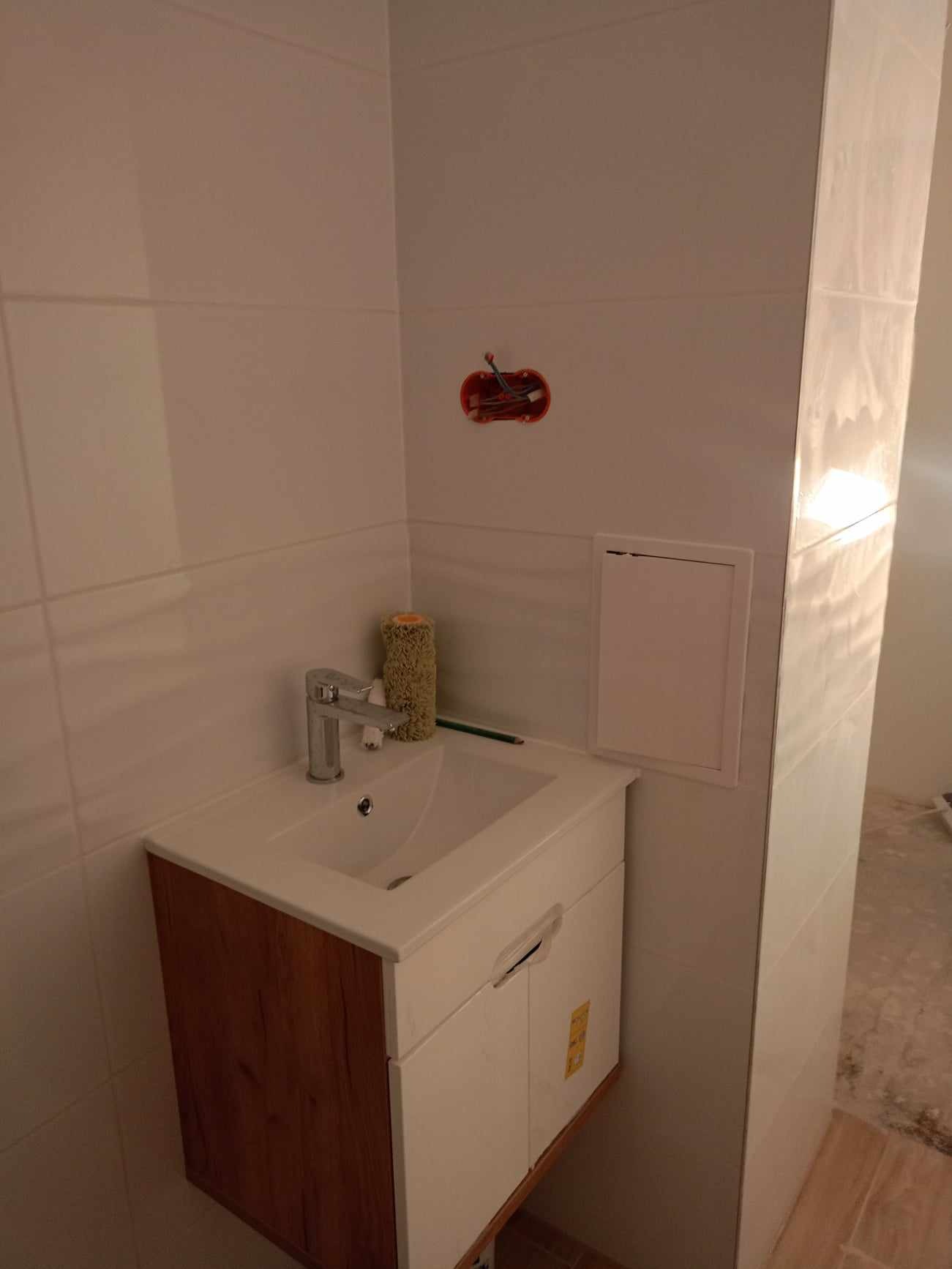 Umywalka z szafką w trakcie montażu w łazience wykończonej białymi, prostokątnymi płytkami; widoczne otwarte gniazdko elektryczne, wałek do malowania i ołówek.