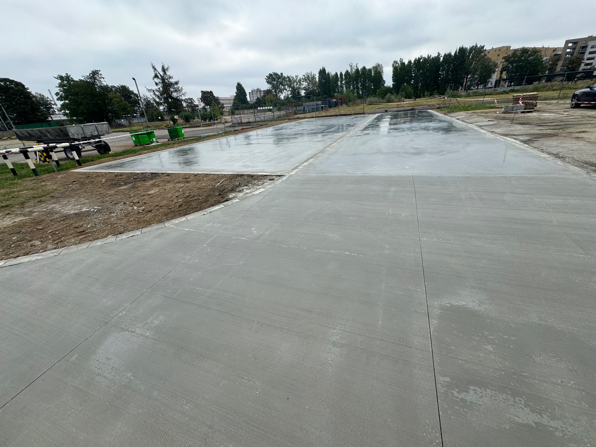 Świeżo wylana, gładka betonowa powierzchnia na zewnątrz, widoczne ślady po zacieraniu, w tle budynki mieszkalne i drzewa pod pochmurnym niebem.