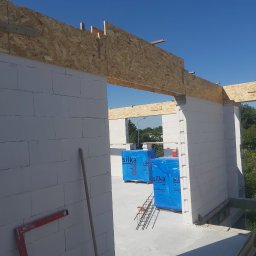 Adkar Bud - Wznoszenie ścian z bloczków silka z drewnianym nadprożem, widoczne paczki z bloczkami w niebieskiej folii, poziomica i łata murarska przy ścianie.