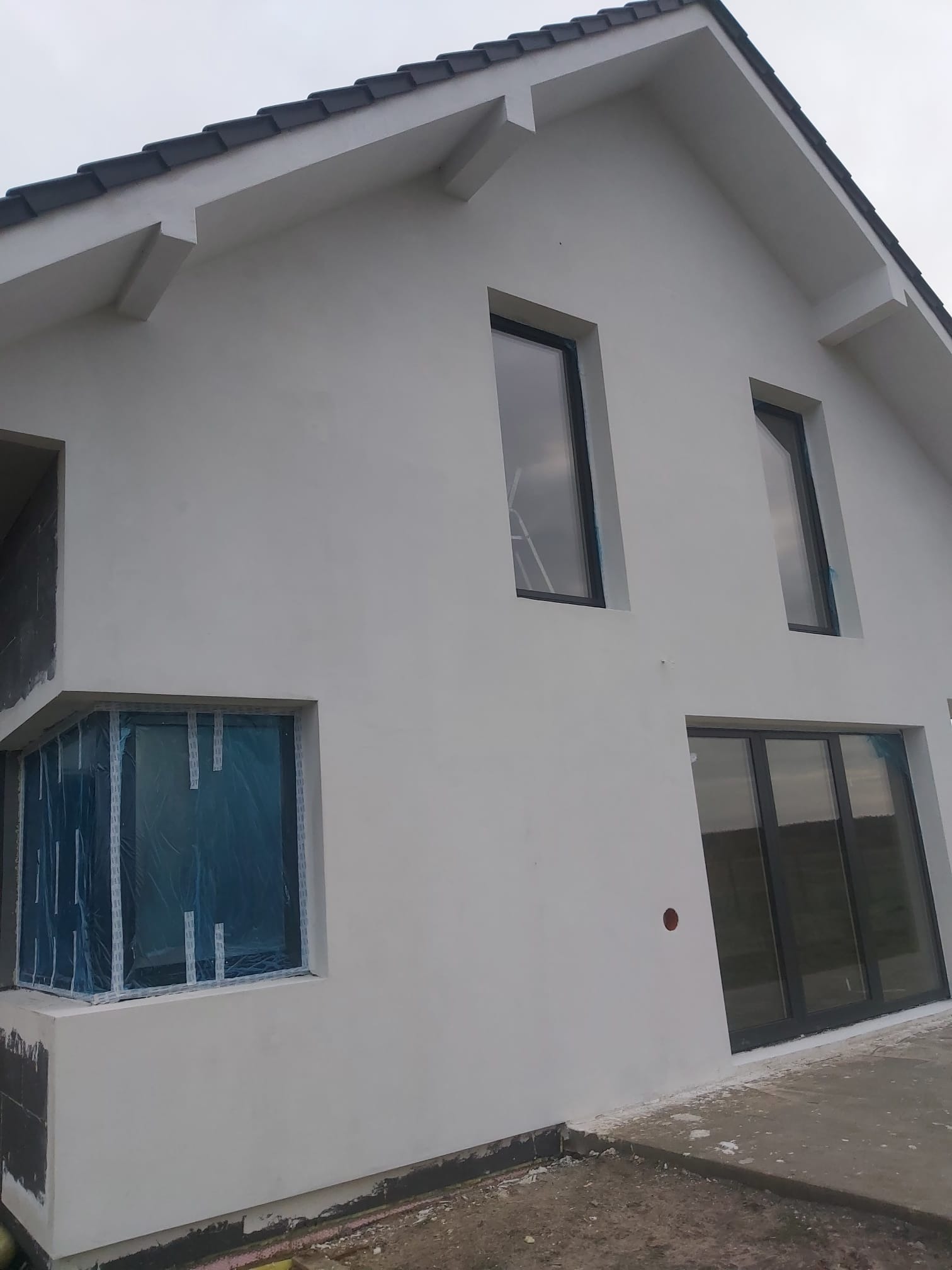 Nowoczesny dom jednorodzinny z szarą elewacją, okna zabezpieczone folią ochronną, widoczne prace wykończeniowe.