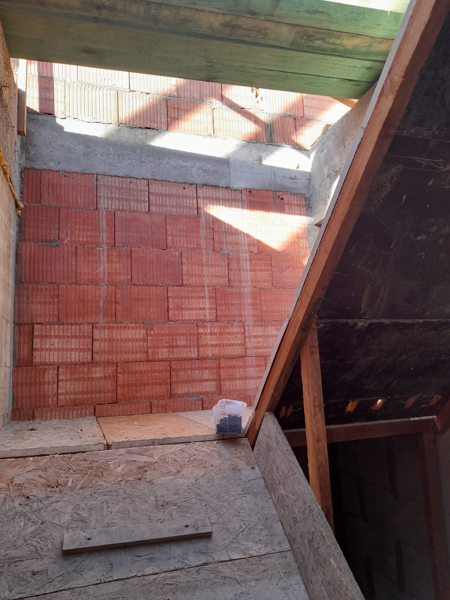 Wnętrze poddasza w trakcie prac: murowana ściana z czerwonej cegły, częściowo otynkowana, widoczne drewniane elementy konstrukcji dachu i płyty OSB, pudełko z wkrętami leży na podłodze.
