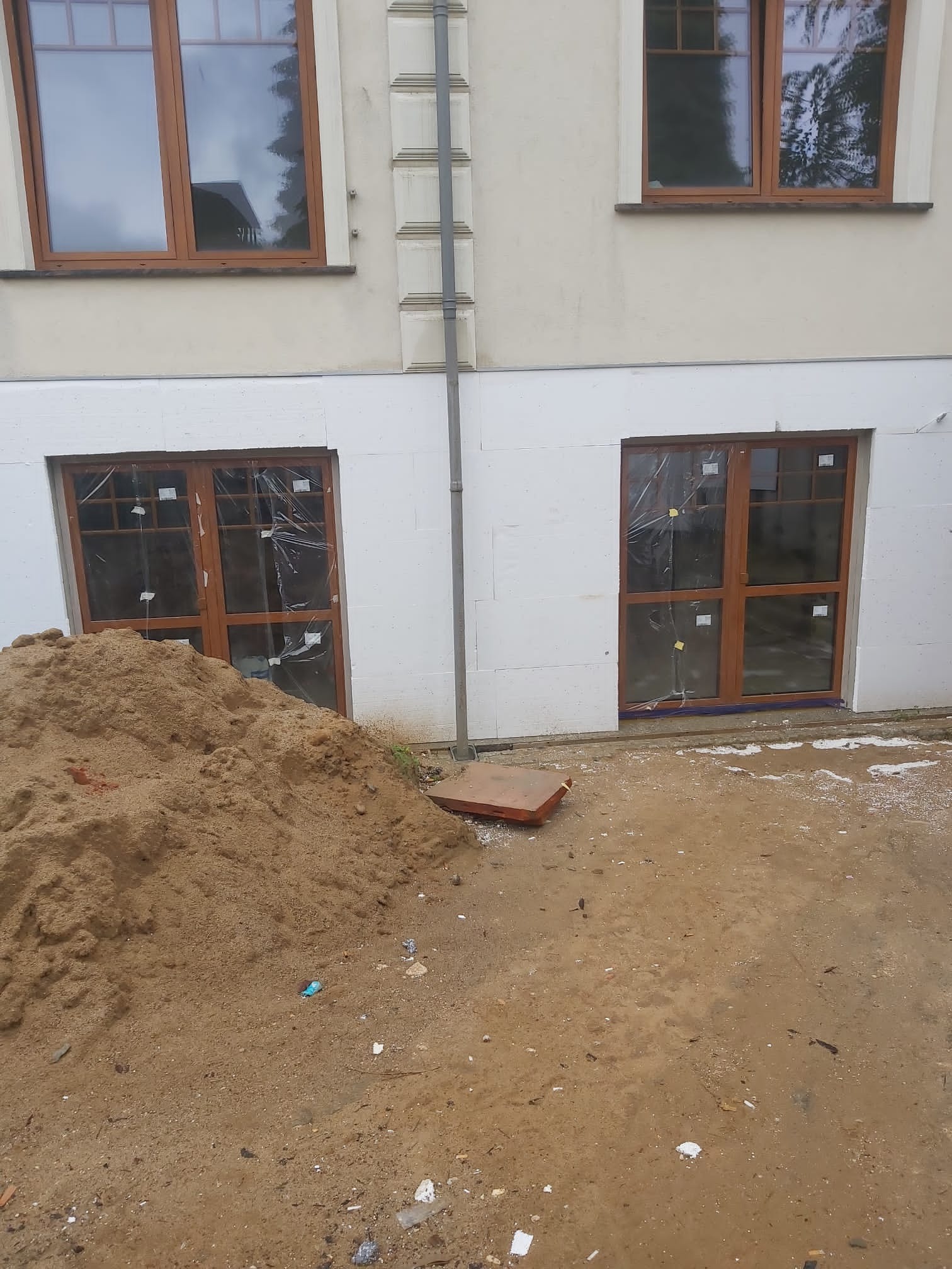 Ocieplanie elewacji styropianem w trakcie prac, widoczne okna zabezpieczone folią i pryzma piasku na ziemi.
