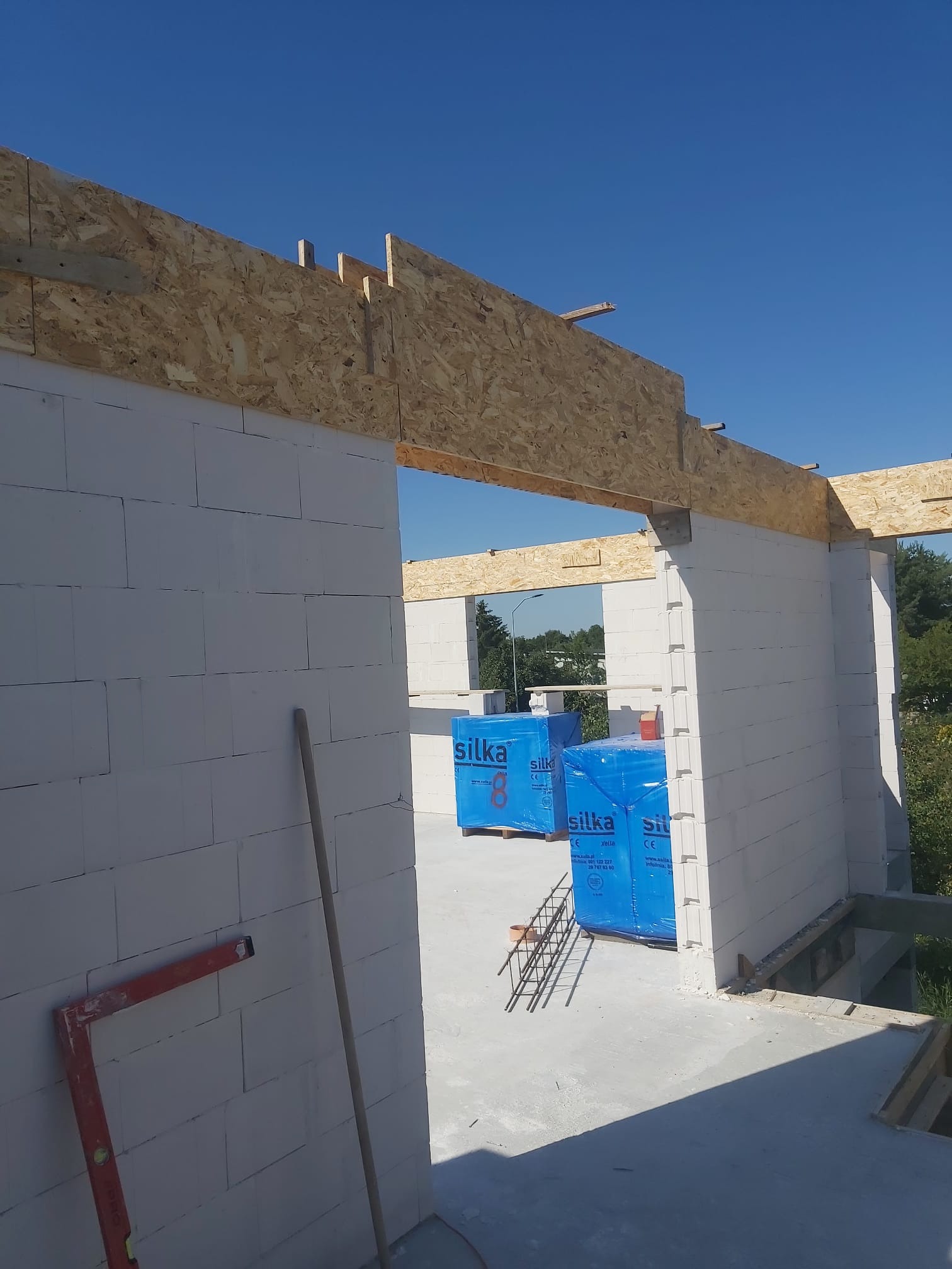 Wznoszenie ścian z bloczków silka z drewnianym nadprożem, widoczne paczki z bloczkami w niebieskiej folii, poziomica i łata murarska przy ścianie.