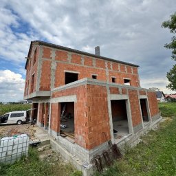 MALAX - Stan surowy otwarty domu z cegły w Wrocławiu, z widocznymi otworami okiennymi i drzwiami, na tle pochmurnego nieba i zieleni. Na pierwszym planie zbiornik na wodę.