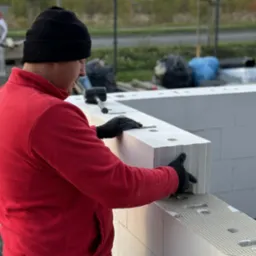 Pracownik w czerwonej bluzie układa białe bloczki na fundamencie. Widoczny fragment ściany i narzędzia. Praca murarska w toku, z dbałością o detale.