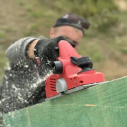 Struganie deski elektrycznym strugiem przez pracownika w rękawicach. Wióry w powietrzu, zielona deska, czerwony strug.