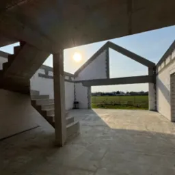 Surowy stan domu z betonu komórkowego w słoneczny dzień: widok na schody, ściany i otwartą przestrzeń z widokiem na zielone pola. Konstrukcja w trakcie budowy.