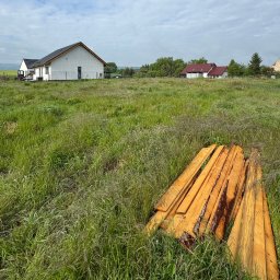 MALAX - Dom w trakcie budowy z ocieploną elewacją w tle, na pierwszym planie stos drewna na zarośniętej działce. Krajobraz wiejski, pochmurne niebo.