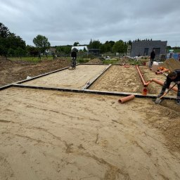 MALAX - Przygotowanie terenu pod fundamenty: ubijanie piasku wewnątrz szalunku, układanie rur kanalizacyjnych. W tle widoczny budynek w trakcie budowy.