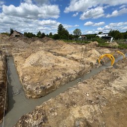 MALAX - Zalane betonem fundamenty w wykopie, widoczne zbrojenia i żółte rury instalacyjne. Tło to hałdy ziemi i błękitne niebo z chmurami.