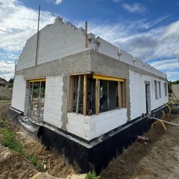 MALAX SPÓŁKA Z OGRANICZONĄ ODPOWIEDZIALNOŚCIĄ - Budowa domu z białych bloczków, widoczne fundamenty zabezpieczone czarną folią, częściowo wzniesione ściany i przygotowane otwory okienne z drewnianą konstrukcją.