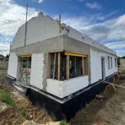 Budowa domu z białych bloczków, widoczne fundamenty zabezpieczone czarną folią, częściowo wzniesione ściany i przygotowane otwory okienne z drewnianą konstrukcją.