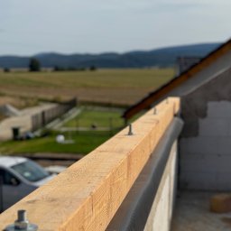 MALAX - Drewniana belka z metalowymi śrubami na murze z bloczków, z widokiem na krajobraz i góry w oddali. Element konstrukcji dachu w trakcie budowy.
