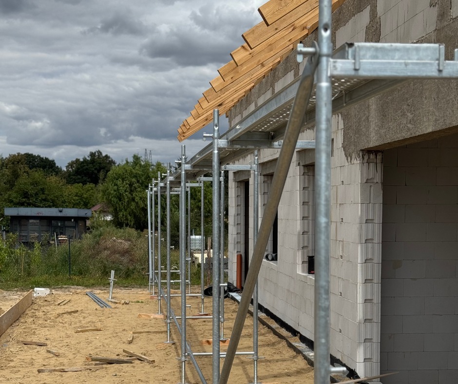 Budowa domu z bloczków: ściany w trakcie murowania, drewniana konstrukcja dachu, rusztowanie. Widok z boku, pochmurne niebo w tle.
