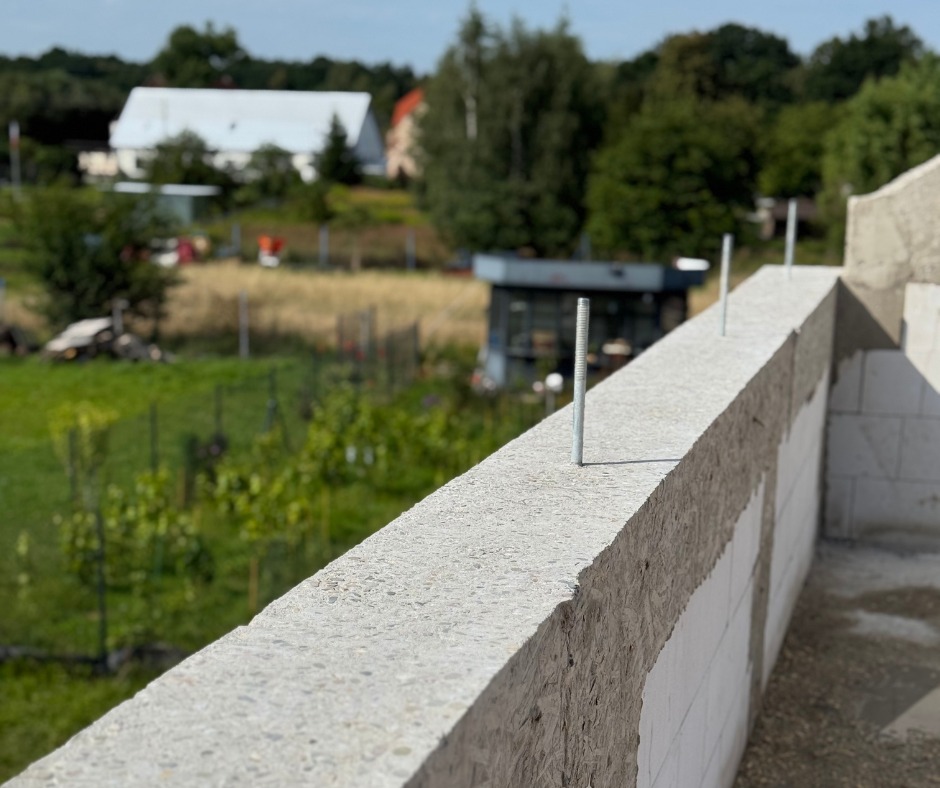 Fragment ściany z betonu komórkowego z wystającymi śrubami kotwiącymi, w tle zieleń i budynki. Widok z góry, perspektywa zbieżna.