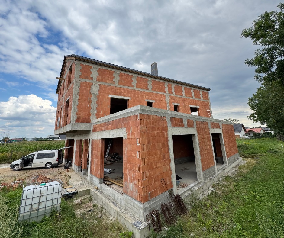 Stan surowy otwarty domu z cegły w Wrocławiu, z widocznymi otworami okiennymi i drzwiami, na tle pochmurnego nieba i zieleni. Na pierwszym planie zbiornik na wodę.