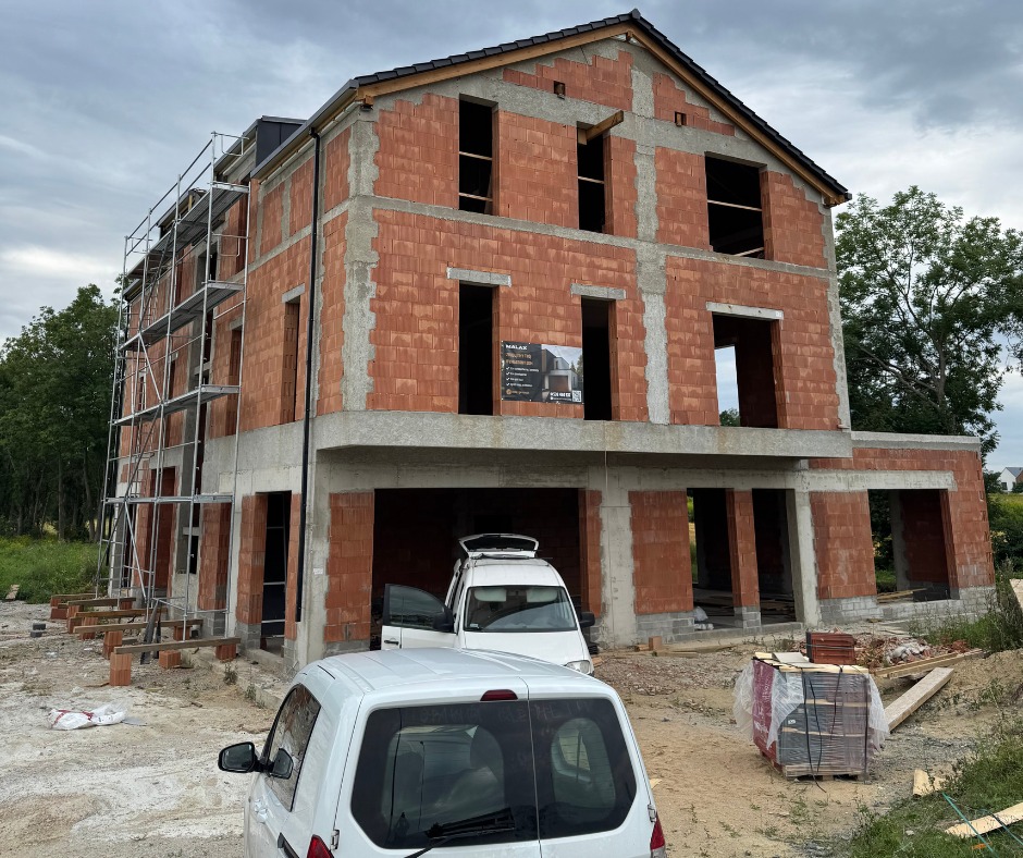 Dwupiętrowy dom w trakcie budowy z czerwonej cegły, z widocznymi rusztowaniami i dwoma samochodami dostawczymi na posesji. Etap murowania ścian.