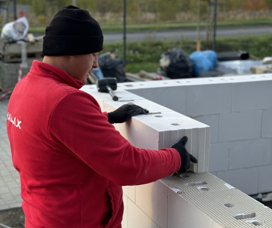 Pracownik w czerwonej bluzie układa białe bloczki na fundamencie. Widoczny fragment ściany i narzędzia. Praca murarska w toku, z dbałością o detale.