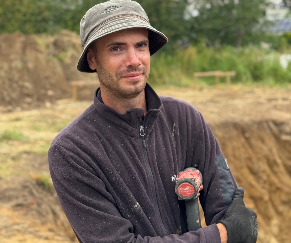 Uśmiechnięty mężczyzna w kapeluszu trzyma wiertarkę udarową na tle wykopu ziemnego, prawdopodobnie pod fundamenty.