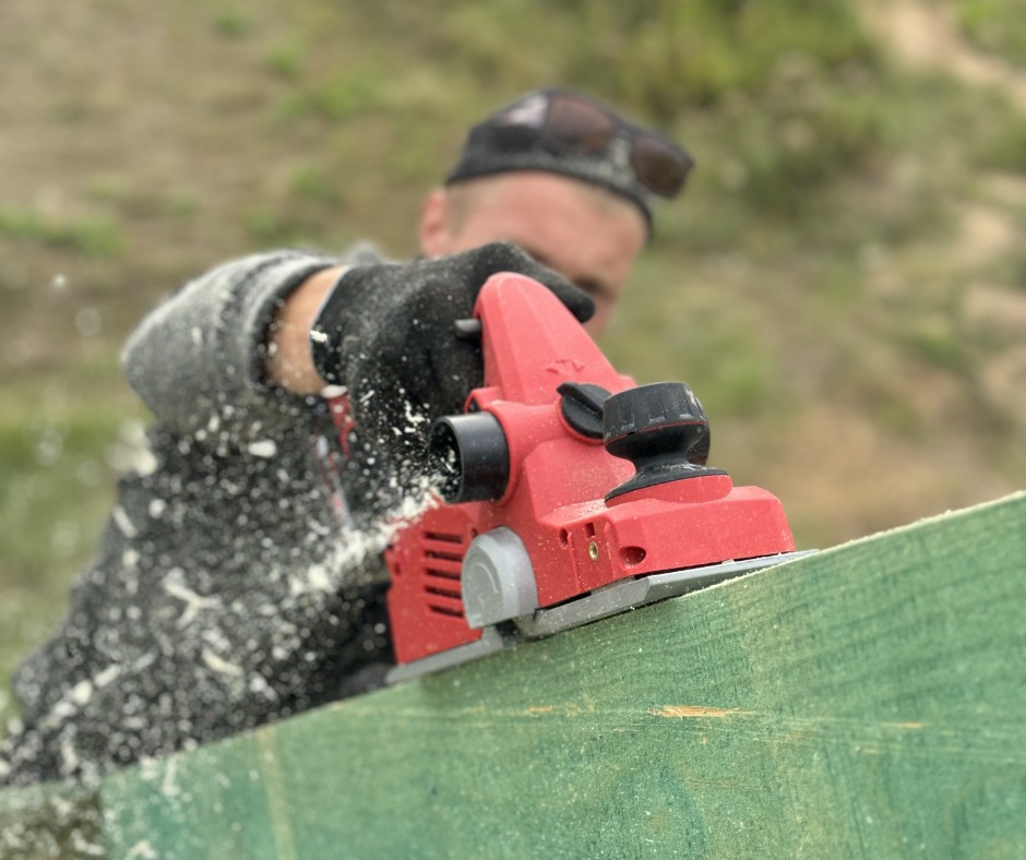 Struganie deski elektrycznym strugiem przez pracownika w rękawicach. Wióry w powietrzu, zielona deska, czerwony strug.