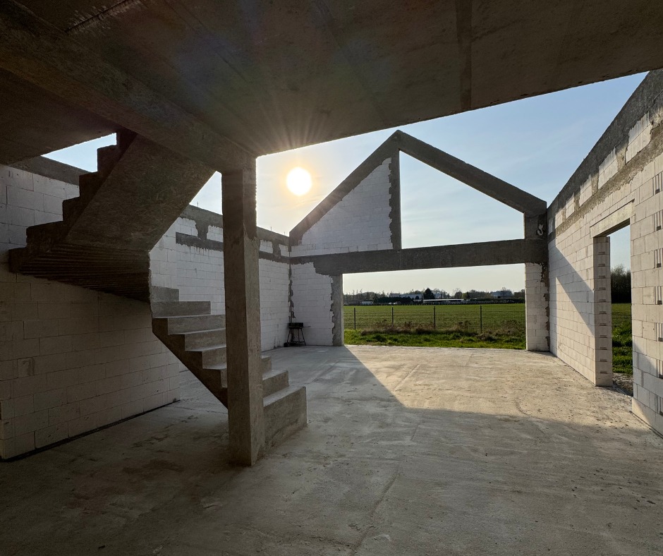 Surowy stan domu z betonu komórkowego w słoneczny dzień: widok na schody, ściany i otwartą przestrzeń z widokiem na zielone pola. Konstrukcja w trakcie budowy.