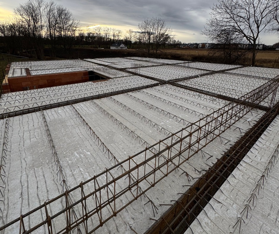 Zbrojony strop Teriva z pustakami, widok z góry na konstrukcję w trakcie budowy domu jednorodzinnego na tle pochmurnego nieba i zabudowy wiejskiej.