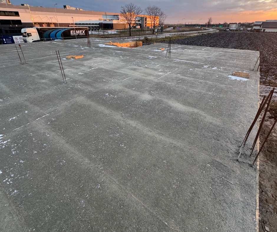 Wylany fundament z wystającymi prętami zbrojeniowymi na tle budynków i nieba o zachodzie słońca. Widoczne ślady po deskowaniu.