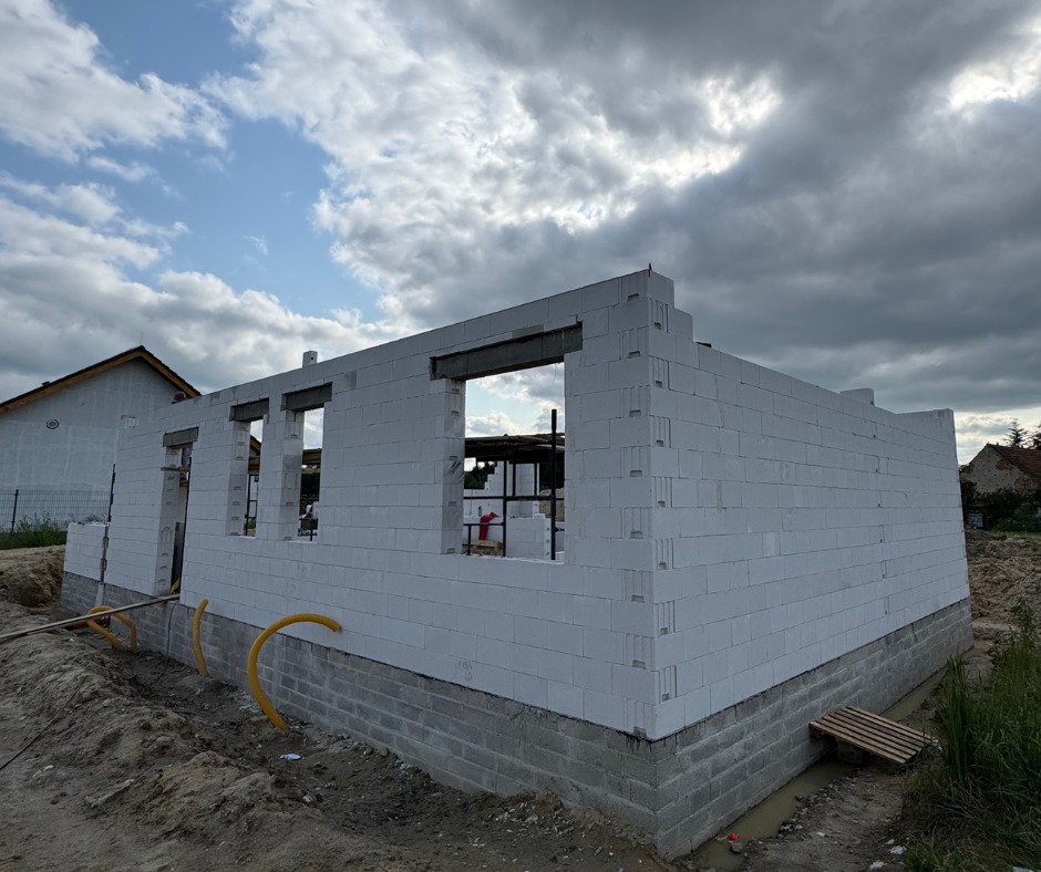 Budowa domu z białych bloczków, widoczne fundamenty i ściany parteru. Na tle pochmurne niebo i częściowo widoczne budynki w oddali. Żółte rury przy fundamencie.