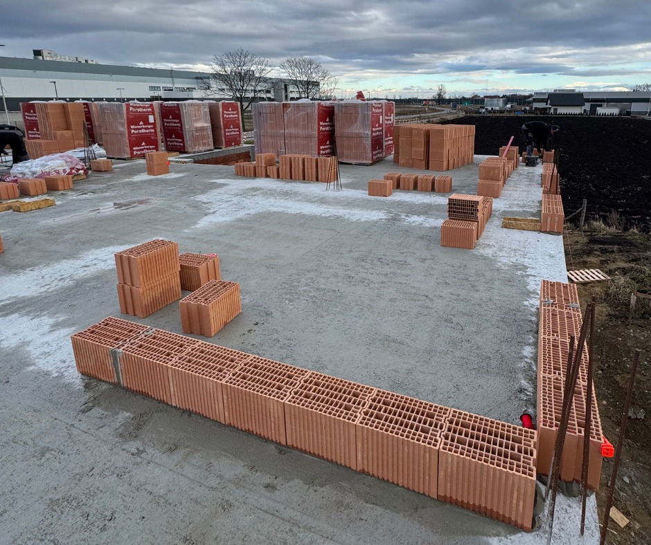 Fundamenty z bloczków ceramicznych Porotherm na wylewce betonowej. Częściowo wymurowane ściany, palety z bloczkami w tle, pochmurne niebo.