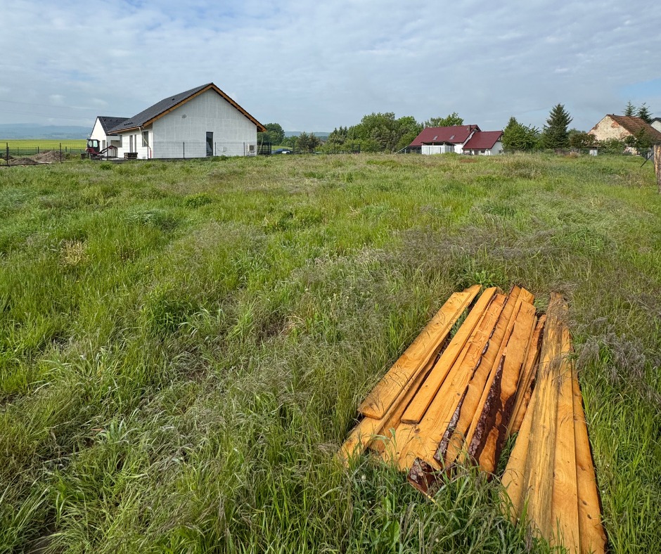 Dom w trakcie budowy z ocieploną elewacją w tle, na pierwszym planie stos drewna na zarośniętej działce. Krajobraz wiejski, pochmurne niebo.