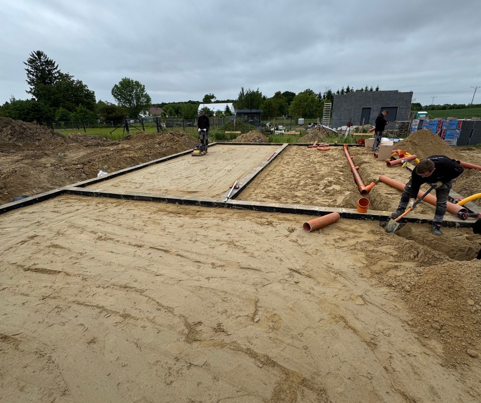 Przygotowanie terenu pod fundamenty: ubijanie piasku wewnątrz szalunku, układanie rur kanalizacyjnych. W tle widoczny budynek w trakcie budowy.