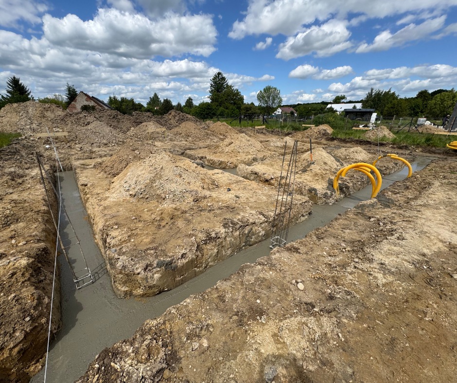 Zalane betonem fundamenty w wykopie, widoczne zbrojenia i żółte rury instalacyjne. Tło to hałdy ziemi i błękitne niebo z chmurami.