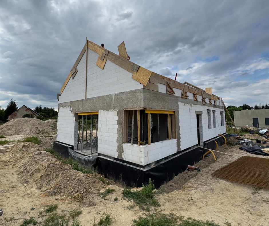 Budowa domu jednorodzinnego w stanie surowym otwartym: ściany z bloczków, widoczne otwory okienne i drzwiowe, przygotowania do dachu, czarna izolacja fundamentów.