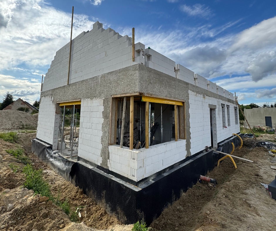 Budowa domu z białych bloczków, widoczne fundamenty zabezpieczone czarną folią, częściowo wzniesione ściany i przygotowane otwory okienne z drewnianą konstrukcją.