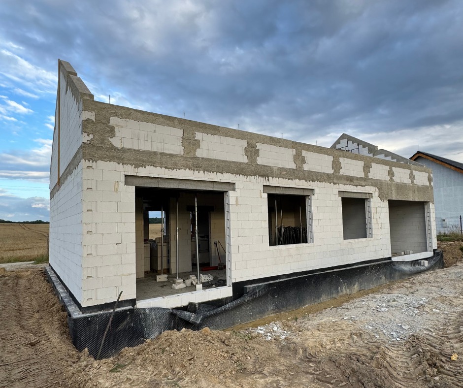 Stan surowy otwarty domu z białego betonu komórkowego, widoczne stalowe podpory, czarna izolacja fundamentów. W tle pole i pochmurne niebo.