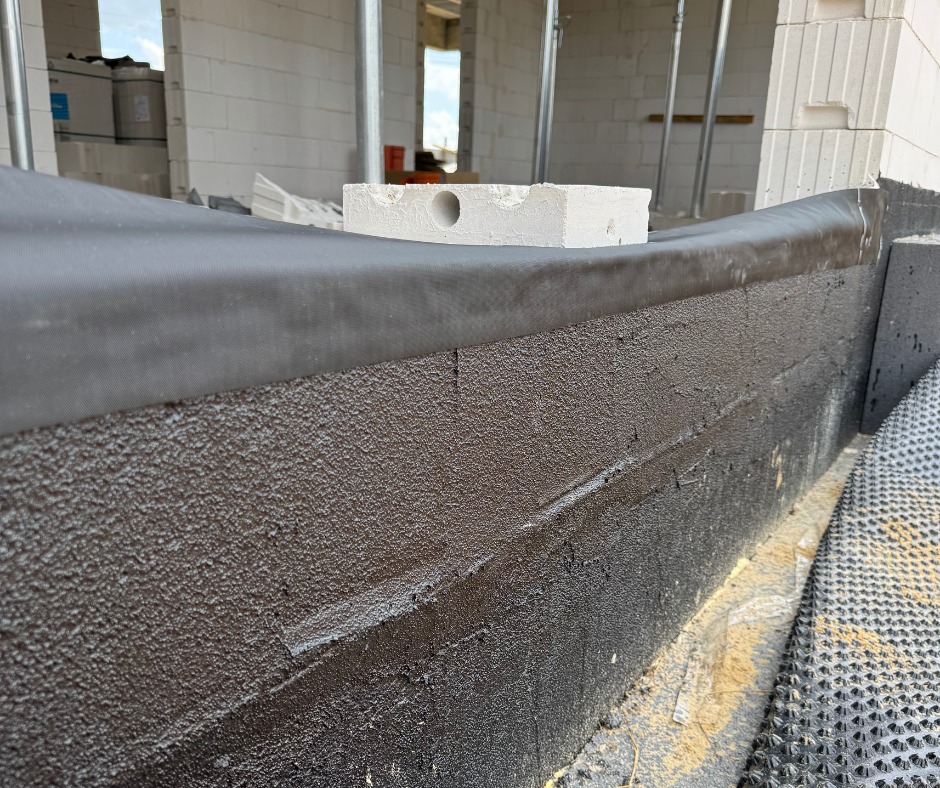 Hydroizolacja fundamentu z użyciem czarnej masy bitumicznej i membrany, widoczny bloczek z betonu komórkowego na górze ściany fundamentowej.