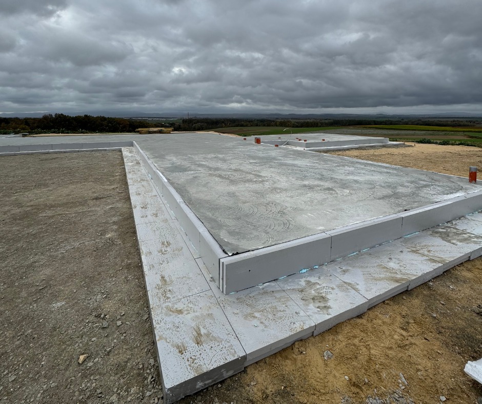 Przygotowany fundament pod budowę domu. Widoczne szalunki i wylany beton. Na horyzoncie zieleń i pochmurne niebo.