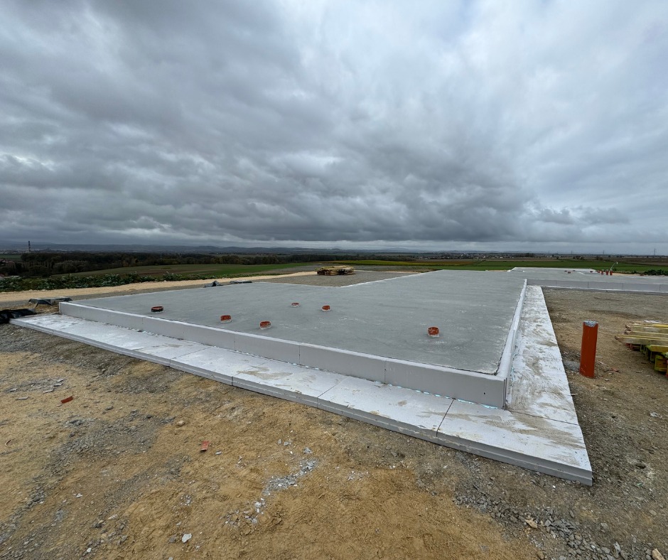 Wylana płyta fundamentowa z izolacją obwodową na budowie domu jednorodzinnego, widok na pola i pochmurne niebo. Etap przygotowania pod dalsze prace budowlane.