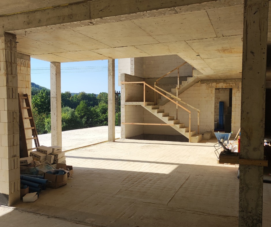 Surowy stan wnętrza domu z betonu komórkowego, z widocznymi schodami i drewnianą drabinką opartą o filar. W tle zieleń drzew i błękit nieba.