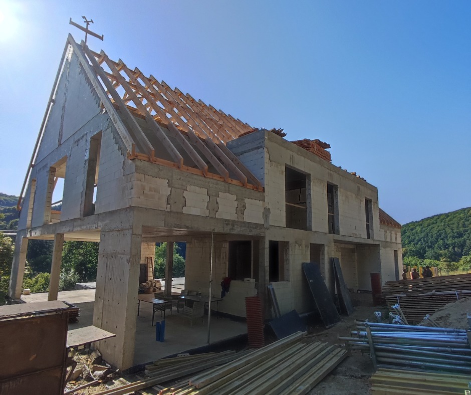 Budowa domu jednorodzinnego w stanie surowym otwartym. Widoczne ściany z betonu komórkowego, więźba dachowa, fundamenty i zarys przyszłego dachu.