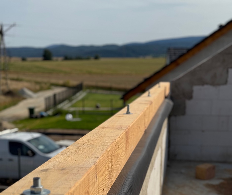 Drewniana belka z metalowymi śrubami na murze z bloczków, z widokiem na krajobraz i góry w oddali. Element konstrukcji dachu w trakcie budowy.