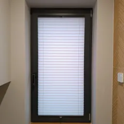 Okno z czarną ramą i zamontowanymi białymi, poziomymi plisami, widok z bliska, drewniany parapet, ściana w kolorze beżowym.