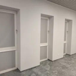 Trzy okna z zamontowanymi roletami wewnętrznymi w biurze z szarą wykładziną i białymi ścianami, widoczne uchwyty okienne.