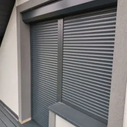 Zewnętrzna roleta antywłamaniowa w kolorze grafitowym, zamontowana na oknie budynku z wykończoną elewacją i drewnianym tarasem.